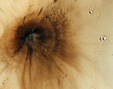 Image of a dark-toned impact crater with streaks radiating from its center, some of which were created when ejecta from the main crater reimpacted the surface and created chains of secondary craters. This impact occurred between December 2003 and November 2005. The main crater itself is only 23 meters across, but the impact event created markings spreading more than a kilometer outward. The interior stands out as blue because the impact excavated a cavity into rocks below the surface that has a different composition than the overlying dust. Some distant dark-toned spots and streaks were created when ejecta from the main crater flew out and reimpacted the surface, producing chains of secondary craters.
